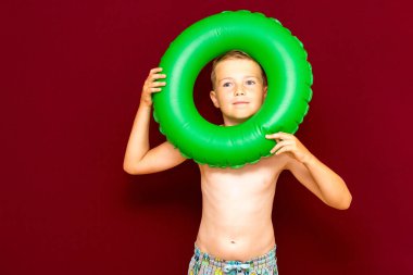 Yüzme çarkındaki delikten çıplak mayo görünümlü beyaz çocuk portresi yeşil şişirilebilir yüzük, yaz tatilinde eğlenen çocuk, stüdyo kırmızı duvar.