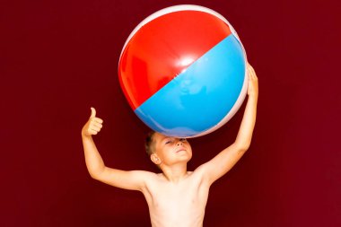 Kırmızı arka planda şişme plaj topu oynayan mutlu beyaz öğrenci başparmağını kaldırıyor. Yaz tatili kavramı