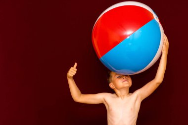 Kırmızı arka planda şişme plaj topu oynayan mutlu beyaz öğrenci başparmağını kaldırıyor. Yaz tatili kavramı