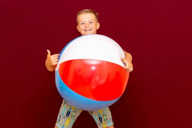 Kırmızı arka planda şişme plaj topu oynayan mutlu beyaz öğrenci başparmağını kaldırdı. Yaz tatili kavramı