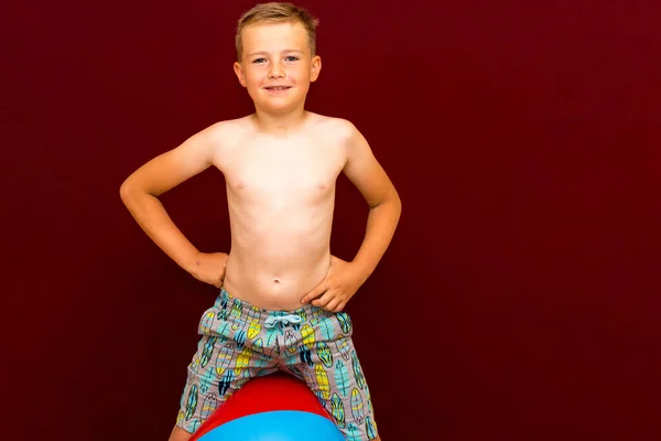 幸せな白人の少年は赤い背景にあるボールインフレータブルビーチボールの上に座っている 夏休みのコンセプト — ストック写真
