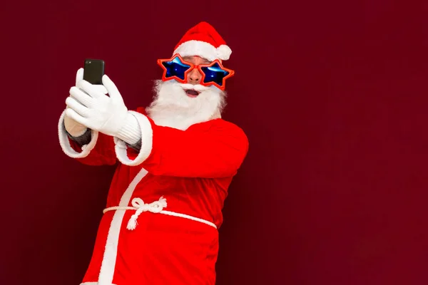 时尚圣诞爪的肖像用手机评论新年帖子 头戴时髦明星眼镜 戴着眼镜 头戴红色背景的帽子 阅读新闻 — 图库照片
