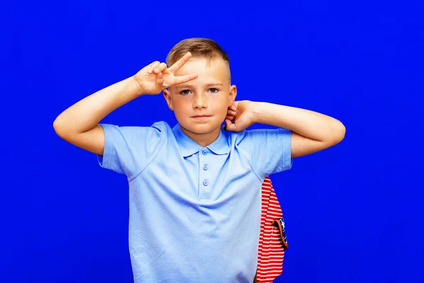 Attractive schoolboy with backpack on the bright blue background shows a sign V. back to school in second grade.