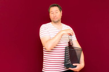 Çizgili tişört giymiş, modaya uygun, tuhaf, orta yaşlı bir adam şarap rengi arka plandaki çöp kutusuna bardak atıyor..