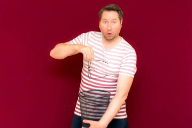 Çizgili tişört giymiş, modaya uygun, orta yaşlı bir adam şarap rengi arka planda çöp kutusuna bardak atıyor.