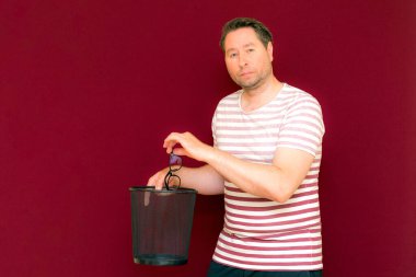 Çizgili tişört giymiş çekici, orta yaşlı bir adam şarap rengi arka plandaki çöp kutusuna bardak atıyor..