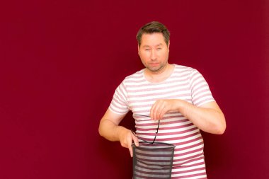 Çizgili tişört giymiş çekici, orta yaşlı bir adam şarap rengi arka plandaki çöp kutusuna bardak atıyor..