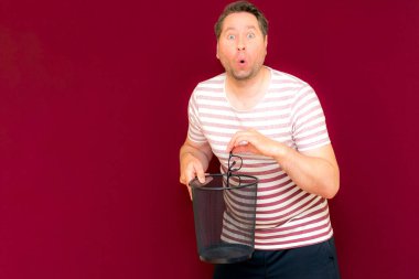 Çizgili tişört giymiş çekici, orta yaşlı bir adam şarap rengi arka plandaki çöp kutusuna bardak atıyor..