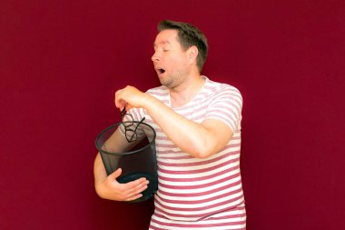 Üzgün, modaya uygun, çekici, orta yaşlı, çizgili tişört giymiş bir adam, şarap rengi arka plandaki çöp kutusuna bardak atıyor..