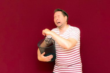 Üzgün, modaya uygun, çekici, orta yaşlı, çizgili tişört giymiş bir adam, şarap rengi arka plandaki çöp kutusuna bardak atıyor..