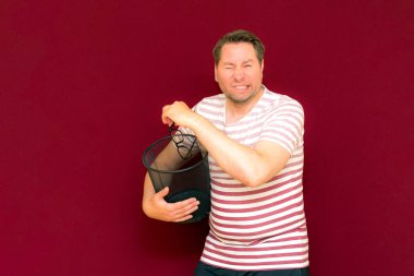 Çizgili tişört giymiş çekici, orta yaşlı bir adam şarap rengi arka plandaki çöp kutusuna bardak atıyor..