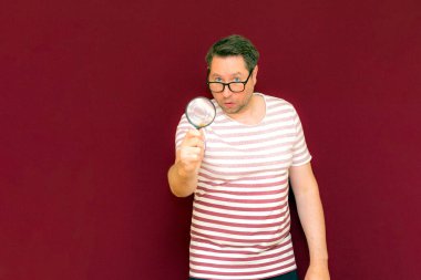 Gözlüklü bir adam çizgili tişört giyiyor. Bir büyüteç alıyor ve içinden bakıyor. Burgundy stüdyo duvarı..