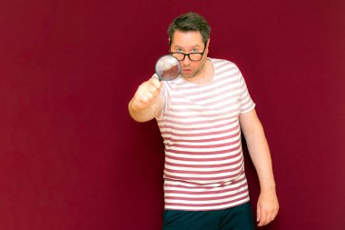 Gözlüklü bir adam çizgili tişört giyiyor. Bir büyüteç alıyor ve içinden bakıyor. Burgundy stüdyo duvarı..