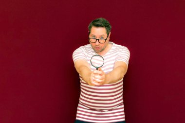 Gözlüklü bir adam çizgili tişört giyiyor. Bir büyüteç alıyor ve içinden bakıyor. Burgundy stüdyo duvarı..