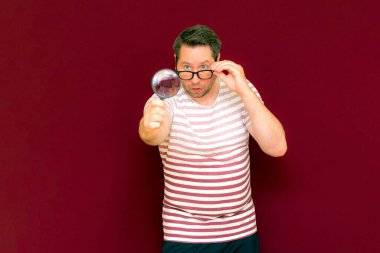Gözlüklü bir adam çizgili tişört giyiyor. Bir büyüteç alıyor ve içinden bakıyor. Burgundy stüdyo duvarı..