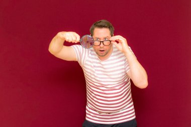Gözlüklü bir adam çizgili tişört giyiyor. Bir büyüteç alıyor ve içinden bakıyor. Burgundy stüdyo duvarı..