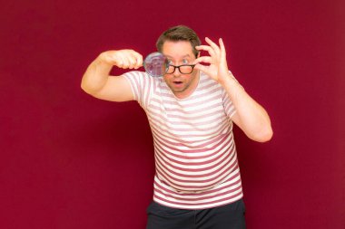 Gözlüklü bir adam çizgili tişört giyiyor. Bir büyüteç alıyor ve içinden bakıyor. Burgundy stüdyo duvarı..