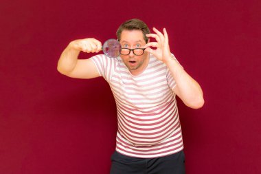 Gözlüklü bir adam çizgili tişört giyer, büyüteçle bakar. Adam şaşırır. Burgundy stüdyo duvarı..