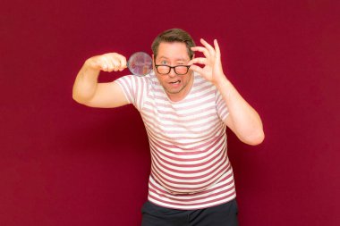 Gözlüklü bir adam çizgili tişört giyiyor. Bir büyüteç alıyor ve içinden bakıyor. Burgundy stüdyo duvarı..
