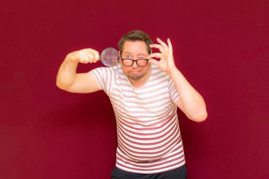 Gözlüklü bir adam çizgili tişört giyiyor. Bir büyüteç alıyor ve içinden bakıyor. Burgundy stüdyo duvarı..