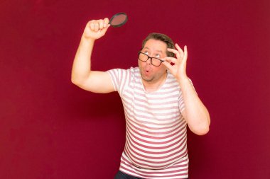 Gözlüklü bir adam çizgili tişört giyiyor. Bir büyüteç alıyor ve içinden bakıyor. Burgundy stüdyo duvarı..