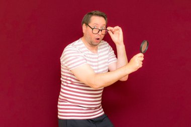 Gözlüklü bir adam çizgili tişört giyer, büyüteçle bakar. Adam şaşırır. Burgundy stüdyo duvarı..