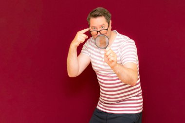 Gözlüklü bir adam çizgili tişört giyiyor. Bir büyüteç alıyor ve içinden bakıyor. Burgundy stüdyo duvarı..