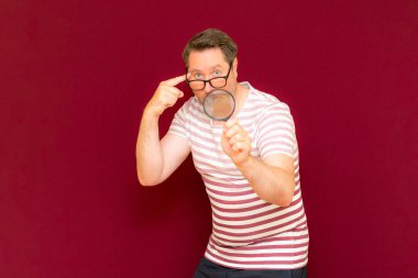 Gözlüklü bir adam çizgili tişört giyiyor. Bir büyüteç alıyor ve içinden bakıyor. Burgundy stüdyo duvarı..