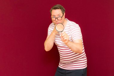 Gözlüklü bir adam çizgili tişört giyiyor. Bir büyüteç alıyor ve içinden bakıyor. Burgundy stüdyo duvarı..