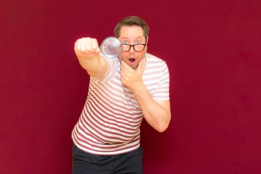 Gözlüklü bir adam çizgili tişört giyer, büyüteçle bakar. Adam şaşırır. Burgundy stüdyo duvarı..