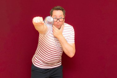 Gözlüklü bir adam çizgili tişört giyiyor. Bir büyüteç alıyor ve içinden bakıyor. Burgundy stüdyo duvarı..