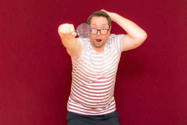 Gözlüklü bir adam çizgili tişört giyer, büyüteçle bakar. Adam şaşırır. Burgundy stüdyo duvarı..