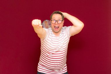 Gözlüklü bir adam çizgili tişört giyer, büyüteçle bakar. Adam şaşırır. Burgundy stüdyo duvarı..