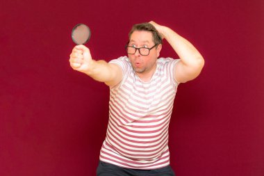 Gözlüklü bir adam çizgili tişört giyer, büyüteçle bakar. Adam şaşırır. Burgundy stüdyo duvarı..