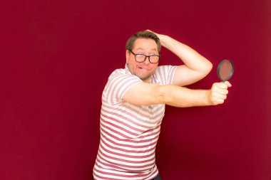 Gözlüklü bir adam çizgili tişört giyiyor. Bir büyüteç alıyor ve içinden bakıyor. Burgundy stüdyo duvarı..