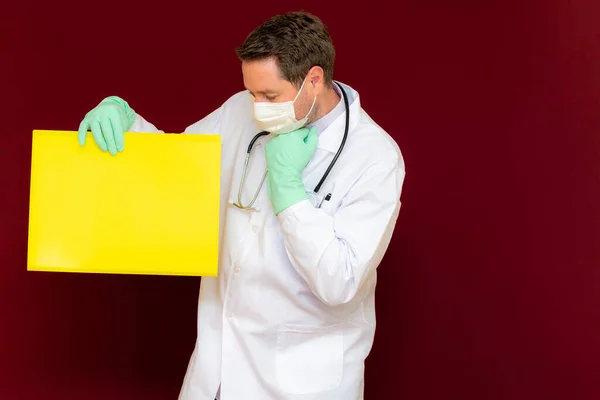 Young handsome doctor wearing medical mask latex gloves and stethoscope on the neck look at yellow blank banner thinks something. Place for your text banner advertisement mock up.Burgundy studio wall.