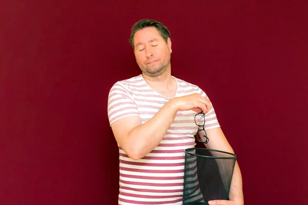 Trendy charming weird middle age man dressed in a striped t shirt throws glasses in the trash bin on a burgundy background.the man closed his eyes