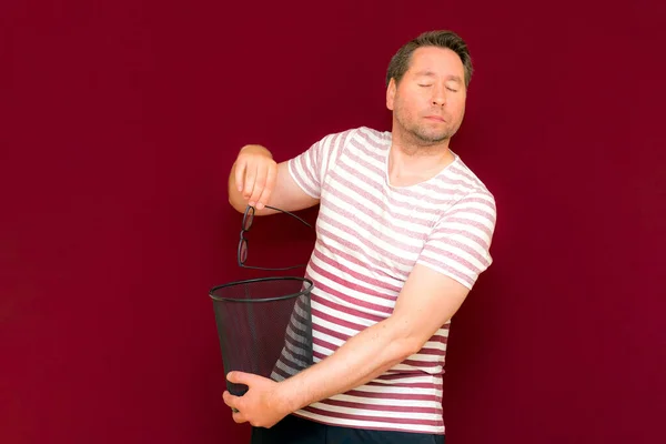 Trendy charming weird middle age closed his eyes man dressed in a striped t shirt throws glasses in the trash bin on a burgundy background.