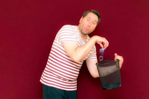 surprised Trendy charming weird middle age man dressed in a striped t shirt throws glasses in the trash bin on a burgundy background.