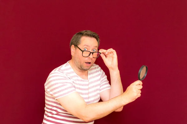 Man with eye glasses wears striped t shirt taking a magnifying glass and looking through it.Man is surprised.Burgundy studio wall.