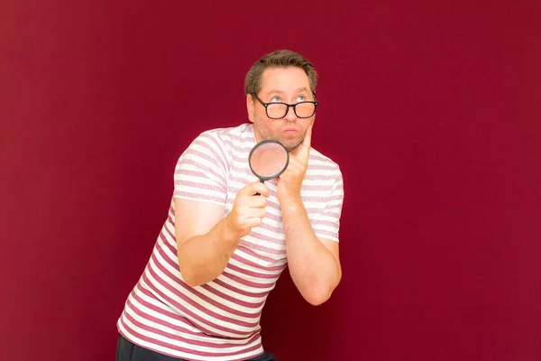 Man with eye glasses wears striped t shirt taking a magnifying glass and looking through it.Burgundy studio wall.
