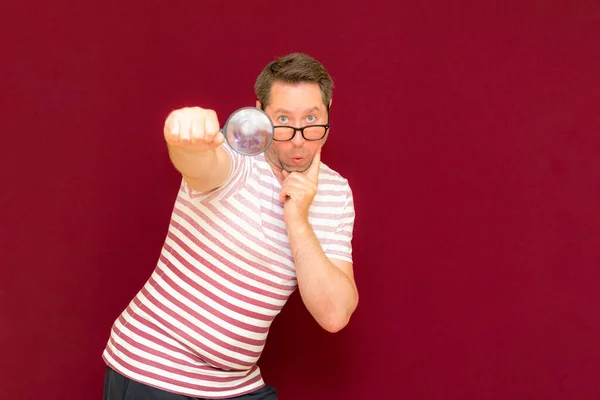Man with eye glasses wears striped t shirt taking a magnifying glass and looking through it.Burgundy studio wall.