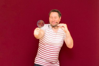 Gözlüklü bir adam çizgili tişört giyiyor. Bir büyüteç alıyor ve içinden bakıyor. Burgundy stüdyo duvarı..