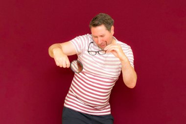 Gözlüklü bir adam çizgili tişört giyiyor. Bir büyüteç alıyor ve içinden bakıyor. Burgundy stüdyo duvarı..