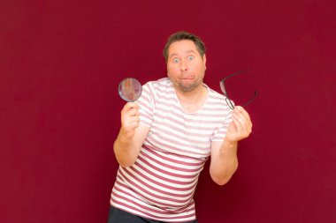 Gözlüklü bir adam çizgili tişört giyiyor. Bir büyüteç alıyor ve içinden bakıyor. Burgundy stüdyo duvarı..