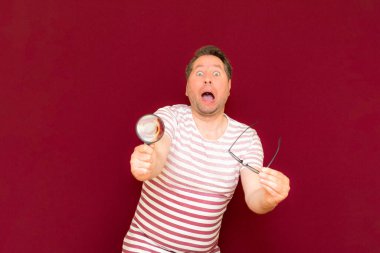 Gözlüklü bir adam çizgili tişört giyiyor. Bir büyüteç alıyor ve içinden bakıyor. Burgundy stüdyo duvarı..