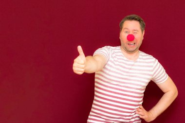 Kendini beğenmiş ve gülümseyen adam kırmızı ya da bordo duvarda baş parmağını gösteriyor. Kırmızı burun günü ya da Nisan 1 'de. Palyaço, eğlence, parti, kutlama, eğlence, eğlence, tatil, mizah konsepti