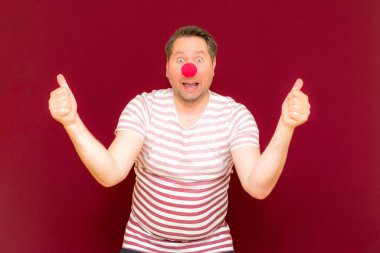 Kendini beğenmiş ve gülümseyen adam kırmızı ya da bordo duvarda baş parmağını gösteriyor. Kırmızı burun günü ya da Nisan 1 'de. Palyaço, eğlence, parti, kutlama, eğlence, eğlence, tatil, mizah konsepti