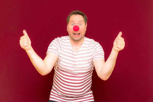 The self-satisfied trendy and smiling man shows thumb up on red or burgundy wall.Red nose day or april fools day. The clown, fun, party, celebration, funny, joy, holiday, humor concept