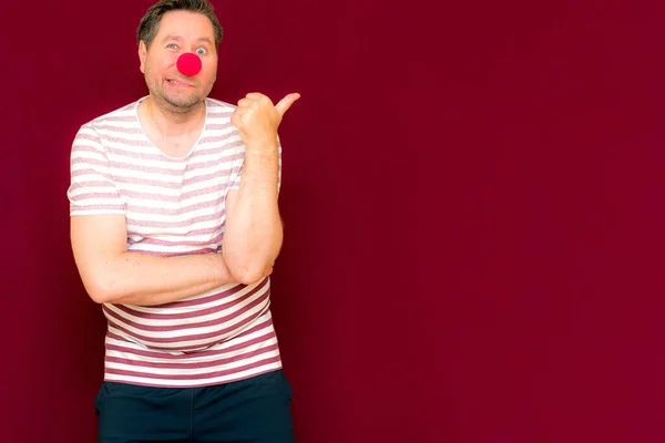 The happy surprised charming trendy and smiling manshows to the side or copy space burgundy wall.Red nose day or april fools day. The clown, fun, party, celebration, funny, joy, holiday,humor concept